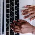 Mani che digitano su una tastiera con schermo di computer in background illuminato da luce d'ufficio