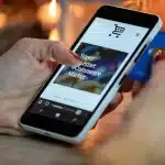Mani di un acquirente che tengono uno smartphone con lista acquisti vicino a una bandiera americana su tavolo da cucina